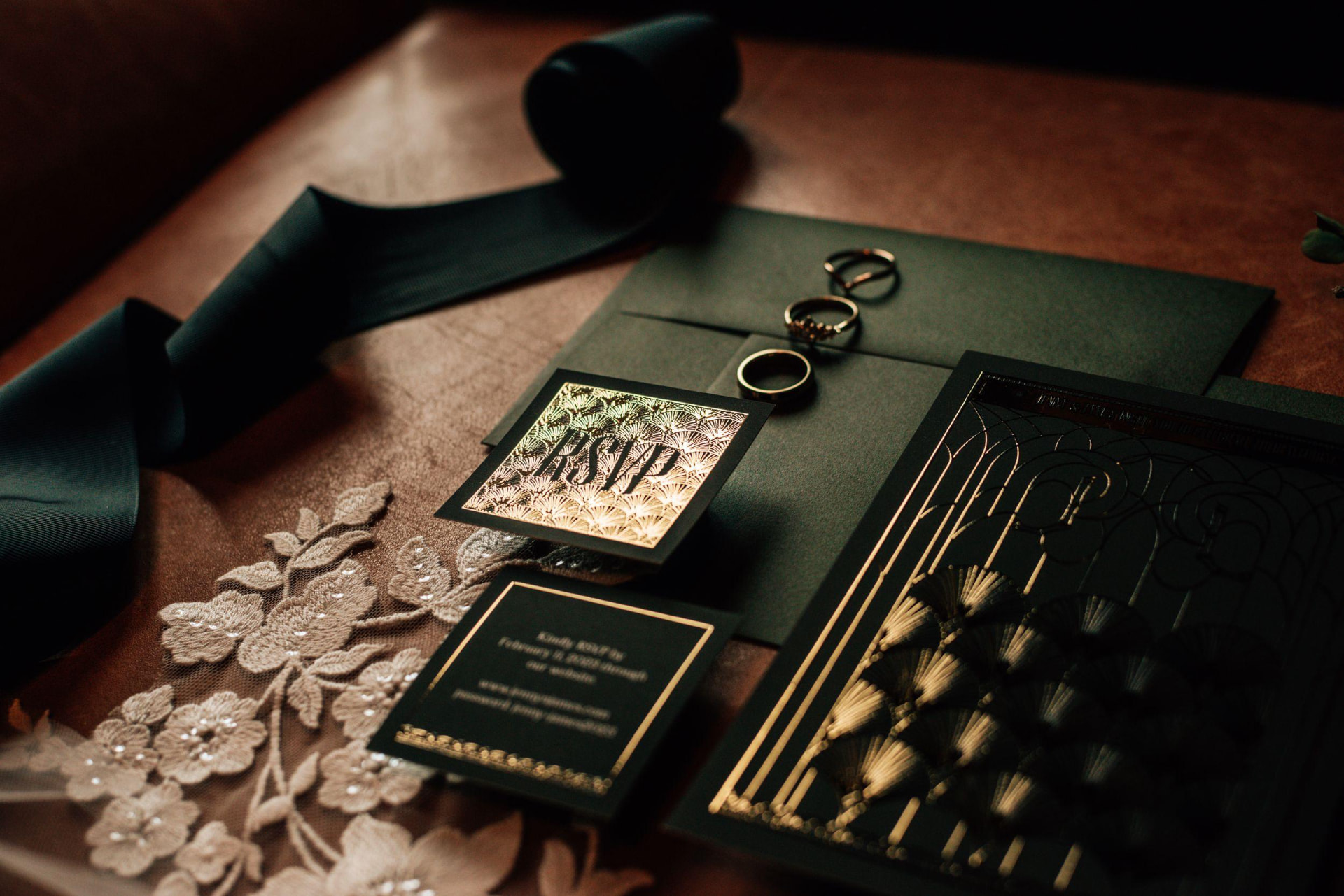 A dark green wedding invitation suite with gold foil, rings and ribbon laid on a leather table